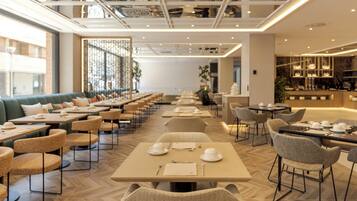 A modern restaurant with long tables and cushioned seating, featuring a herringbone floor pattern and a high ceiling with recessed lighting.