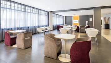 Un hall de hotel con mesas y sillas redondas, un ventanal con cortinas translúcidas y un cartel de ILUNION Hotels.