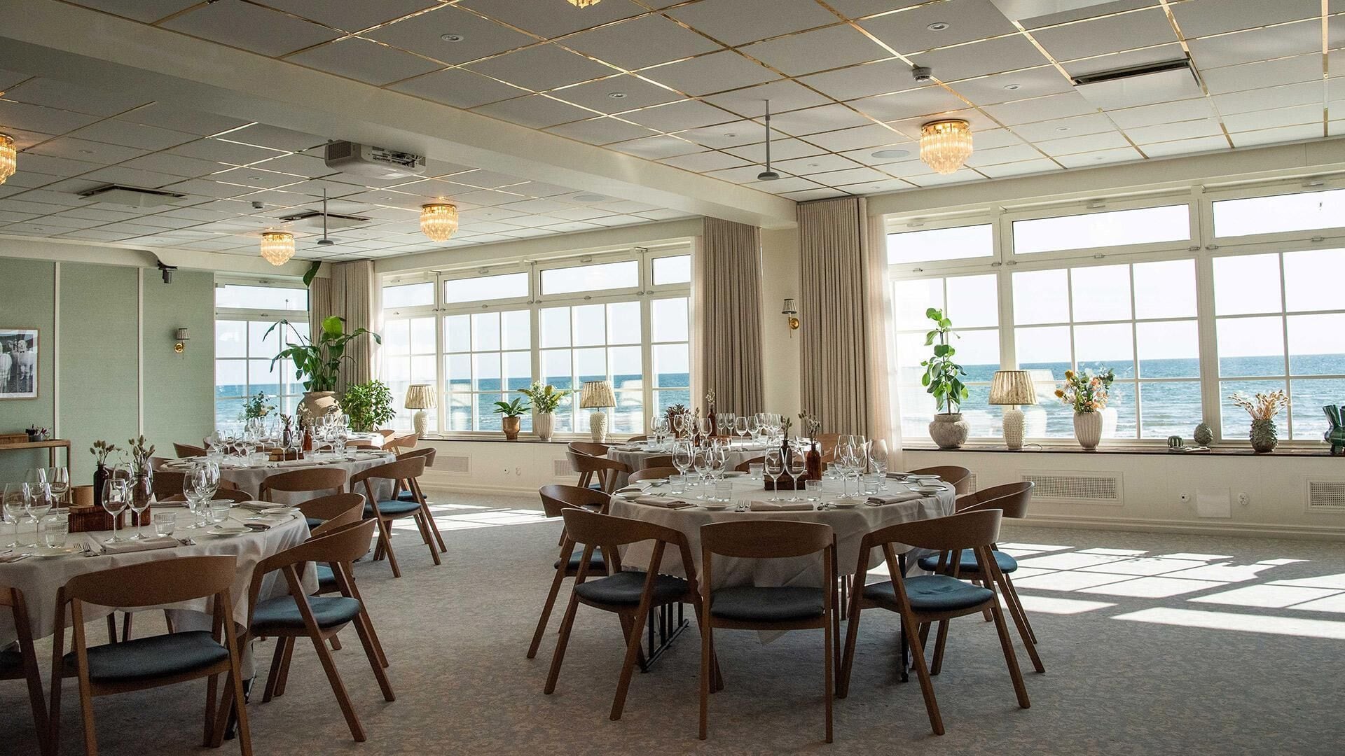 A dining area with tables set for a meal, large windows with a view of the sea, and ceiling lights.
