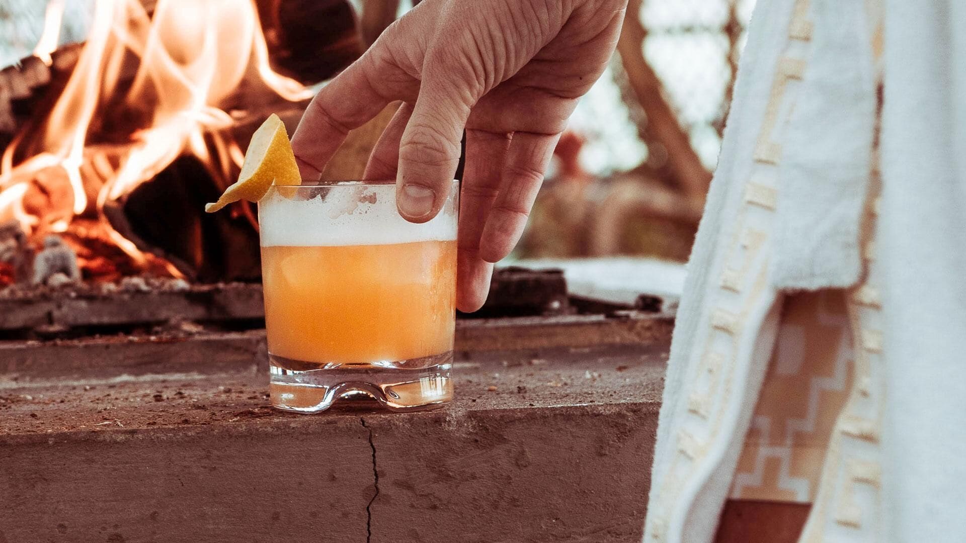 Eine Hand hält ein Glas mit einem schichtartigen Getränk, platziert auf einer Holzoberfläche in der Nähe eines Feuers.