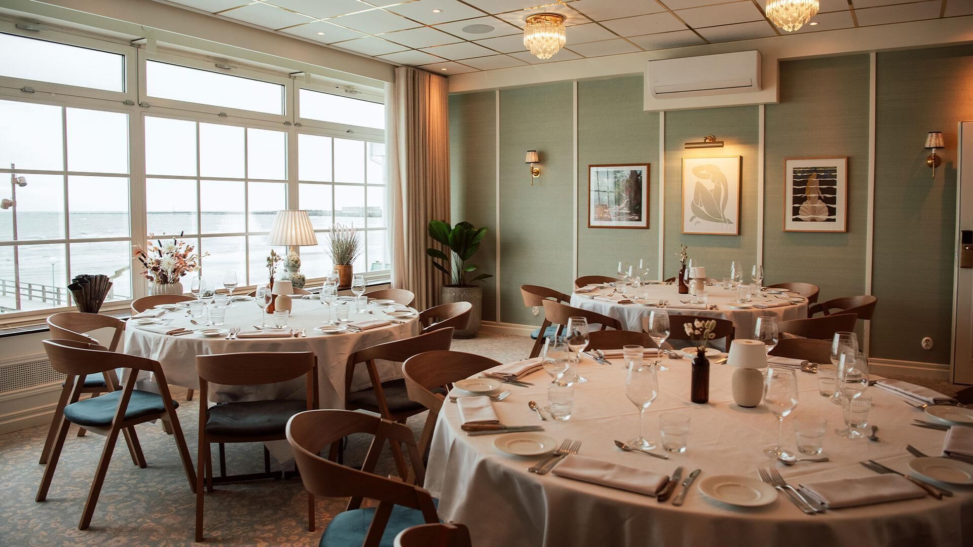 Un espace repas avec des tables rondes disposées pour accueillir plusieurs convives, de grandes fenêtres offrant une vue sur la mer et des luminaires fixés au mur.