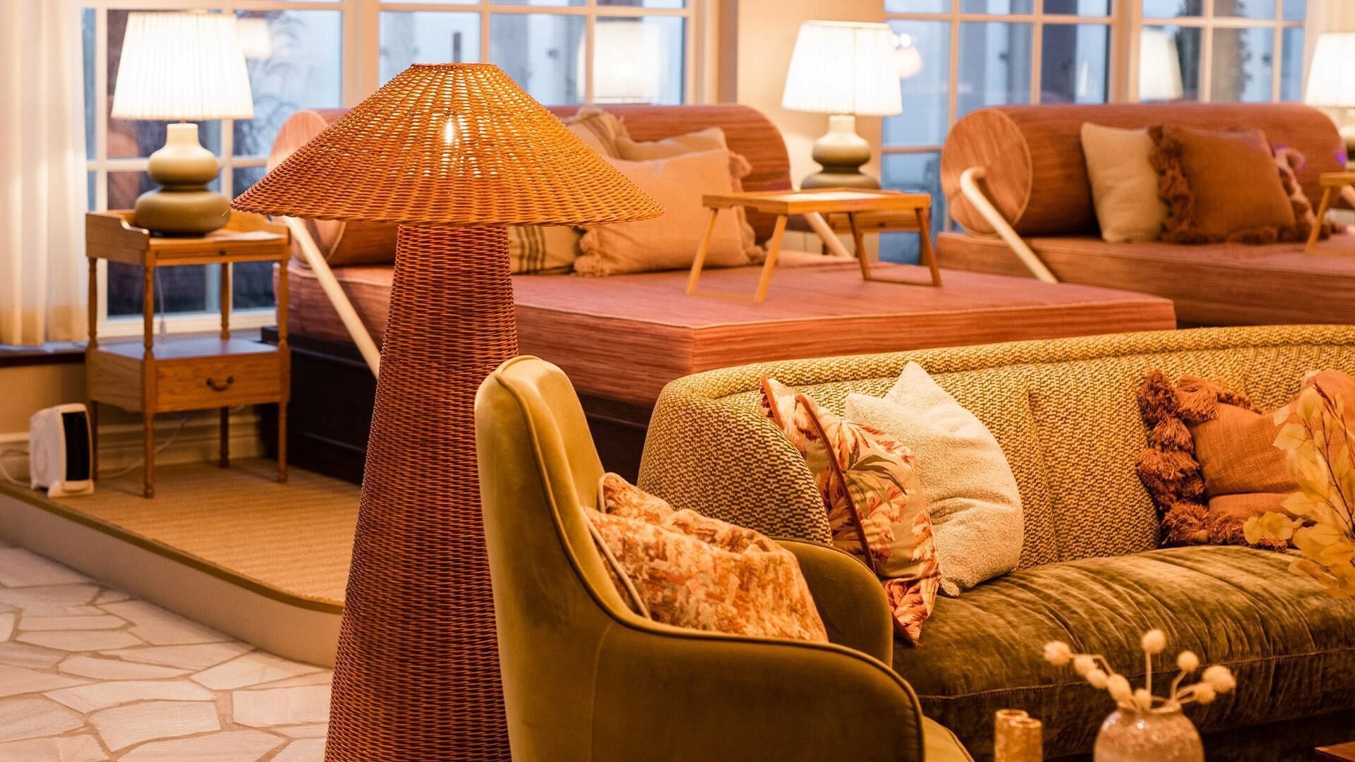 Un salón acogedor con una lámpara de pie trenzada, un sillón verde y un sofá con cojines estampados. Hay dos mesitas de noche con lámparas, y una ventana con vistas al exterior.