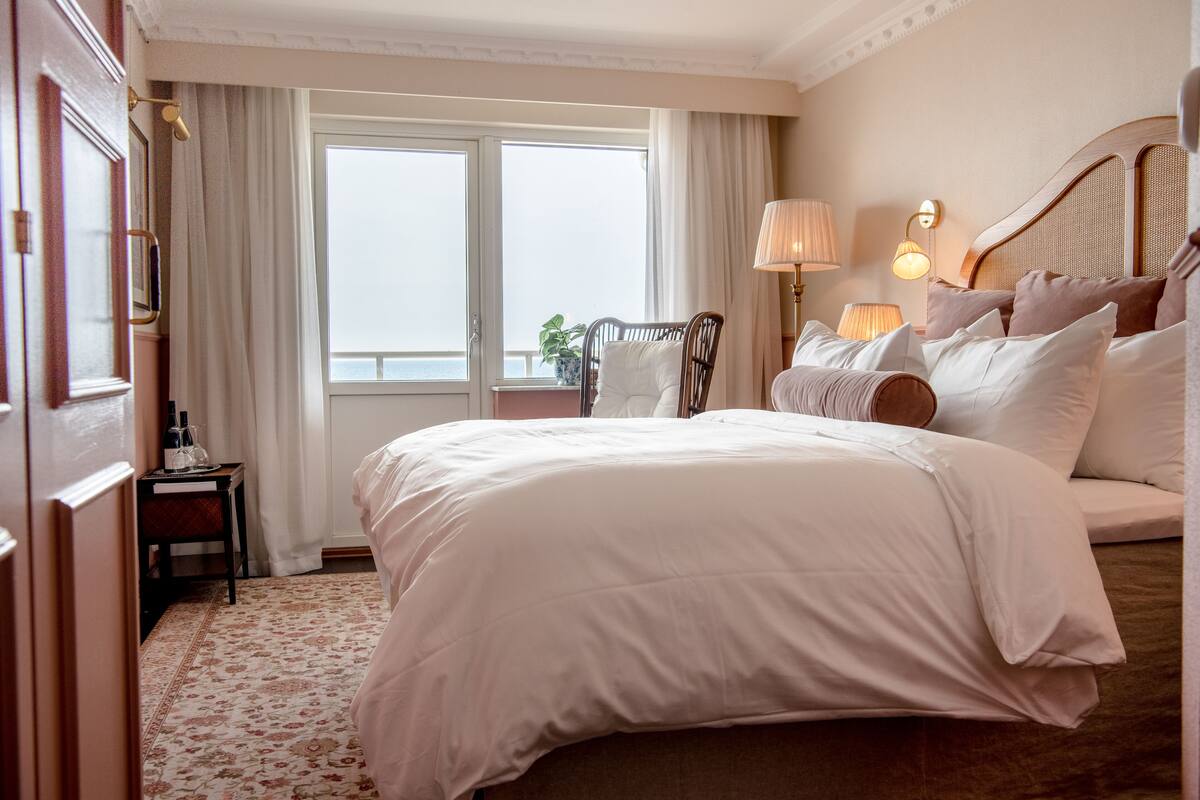 A hotel room with a large bed, a wooden headboard, a nightstand, a lamp, and a view of the sea through a window.
