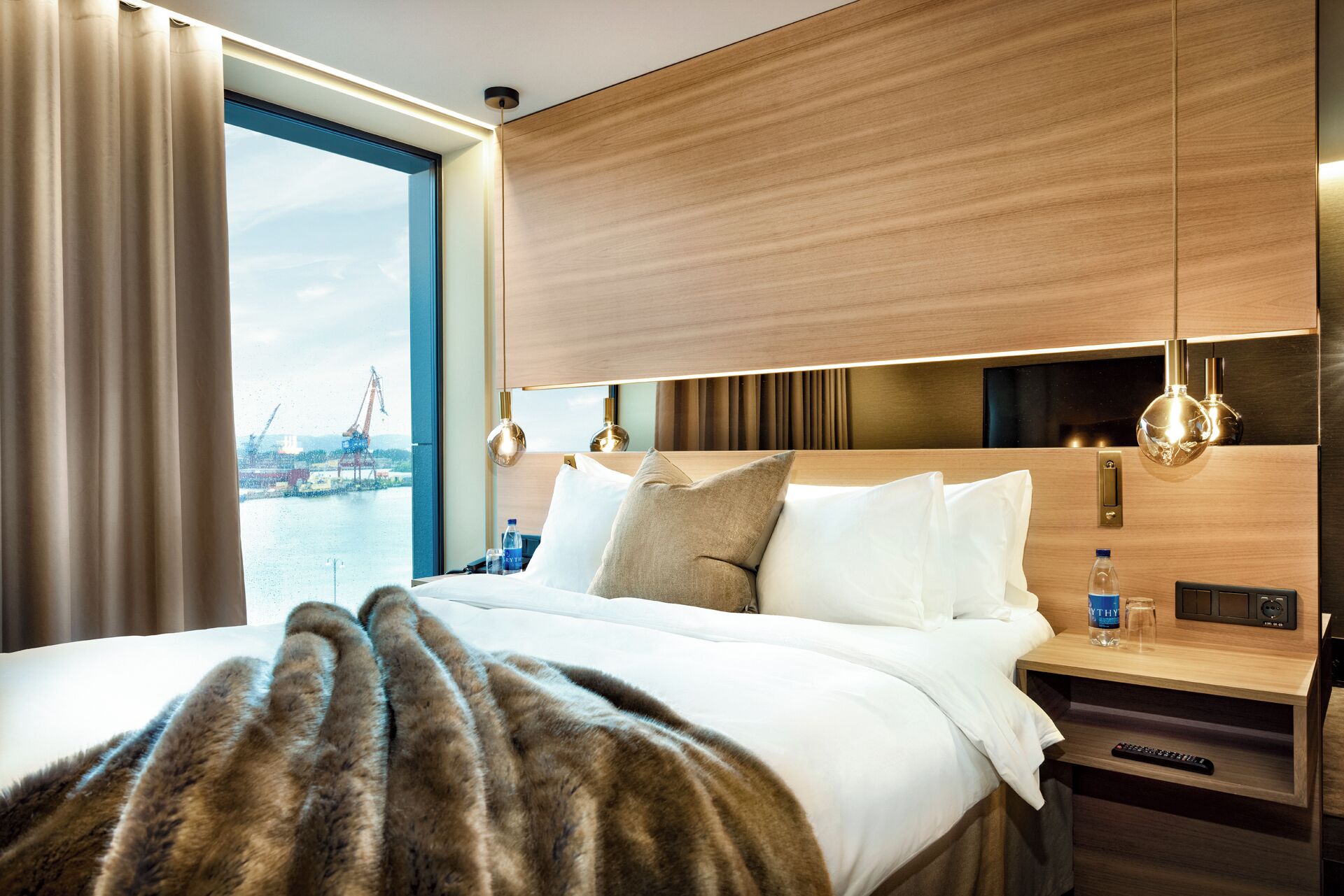 A hotel room with a large bed, a wooden headboard, bedside tables, a nightstand, a lamp, and a view of a body of water with a crane.