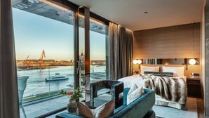 A modern hotel room with a large window offering a view of a river and cityscape, a bed with a fur throw, a seating area with a vase of flowers, and a nightstand with a lamp. - Hotel Riverton (Gothenburg)