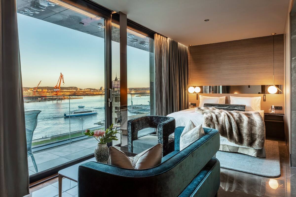 A modern hotel room with a large window offering a view of a river and cityscape, a bed with a fur throw, a seating area with a vase of flowers, and a nightstand with a lamp.