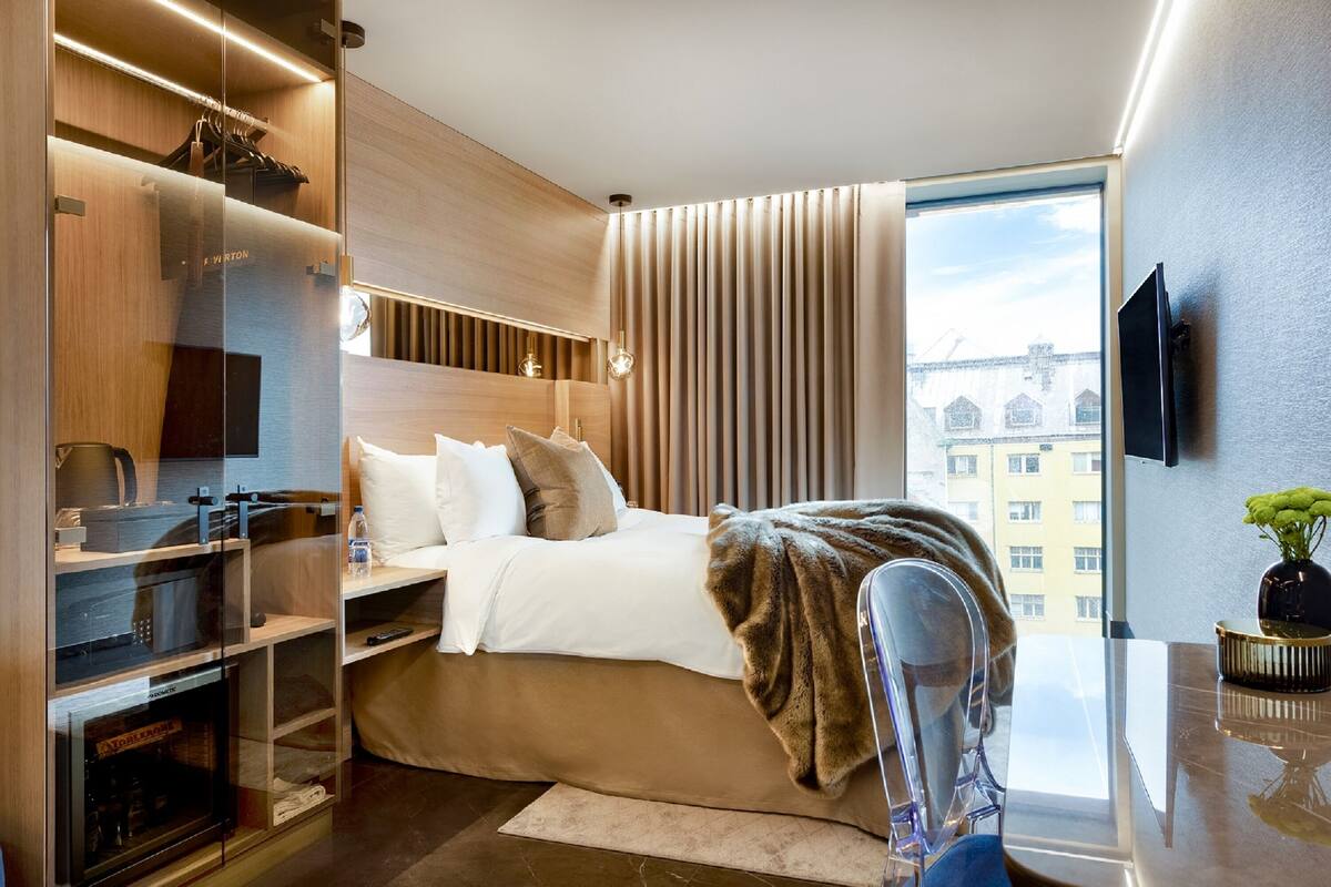 A modern hotel room with a large bed, built-in wardrobe, a TV, and a view of buildings outside.