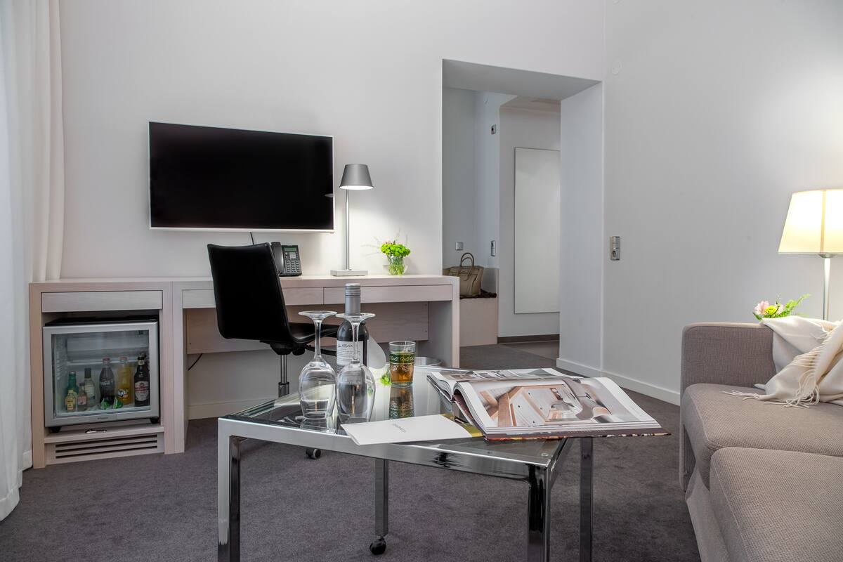 A modern hotel room with a flat-screen TV mounted on the wall, a glass coffee table with magazines, a small refrigerator with beverages, a sofa, and a lamp.