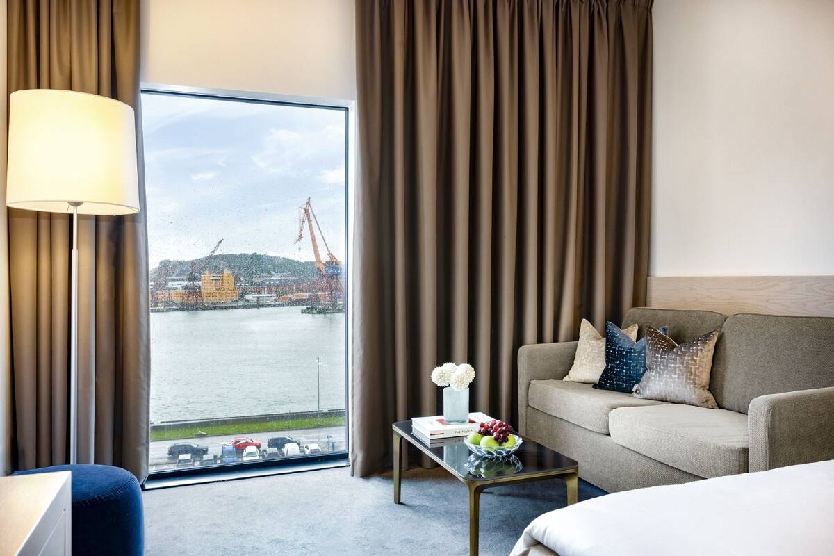 A hotel room with a large window, a sofa, a coffee table with a book and a vase of flowers, and a lamp.
