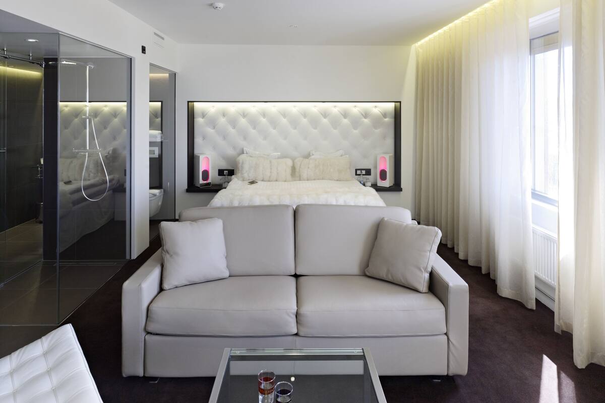 A modern hotel room with a large bed, a sofa, a glass shower enclosure, and a view of the outdoors.