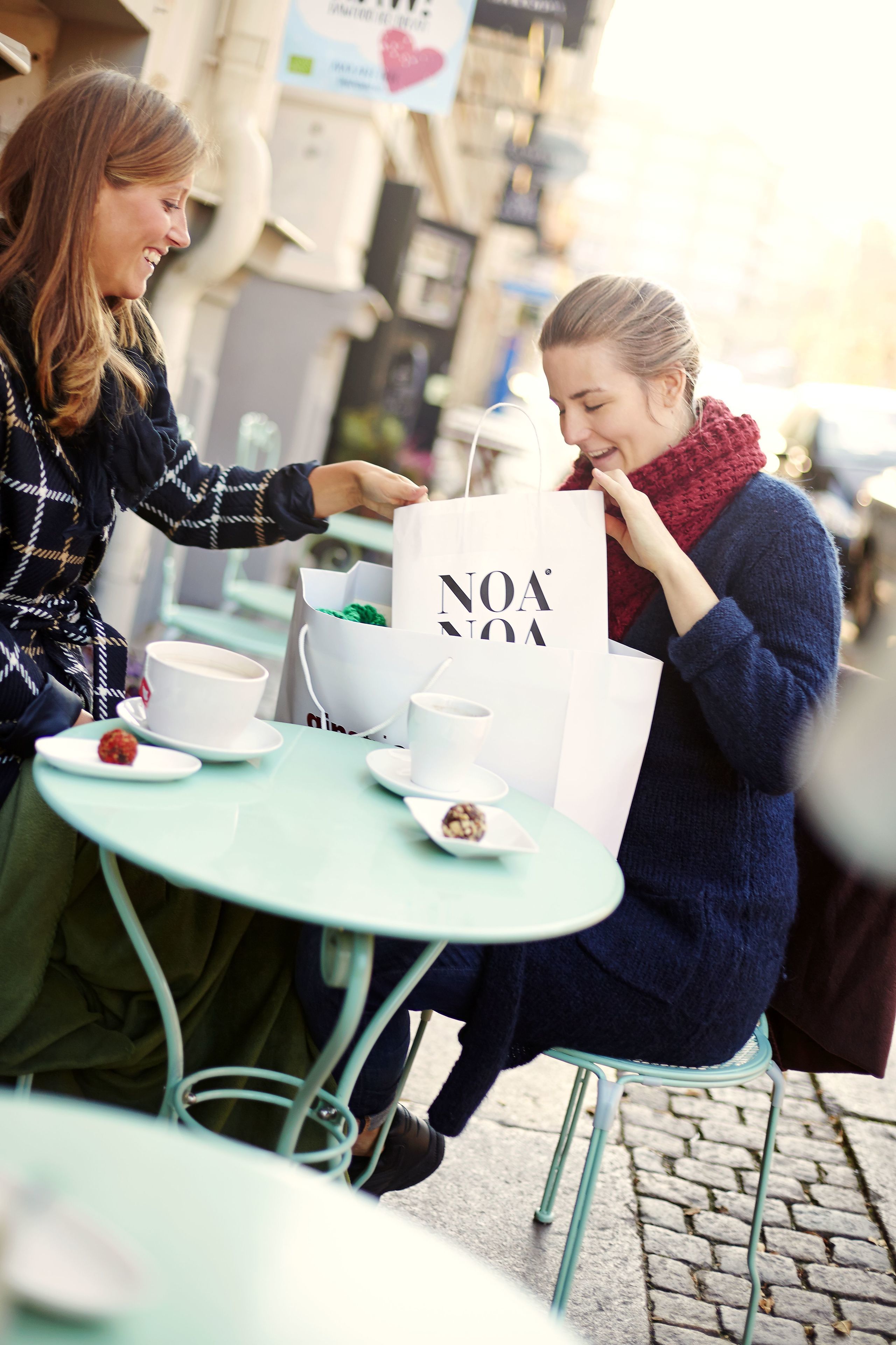 Two people sitting at a table with a cup and saucer, a plate with a strawberry, and a white shopping bag with "NOA" text.