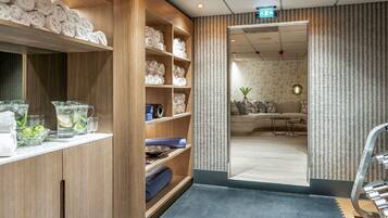 Et hotel lobby med træhylder, der viser håndklæder og toiletartikler, et siddeområde med sofa og bord, samt en væg med et mønster.