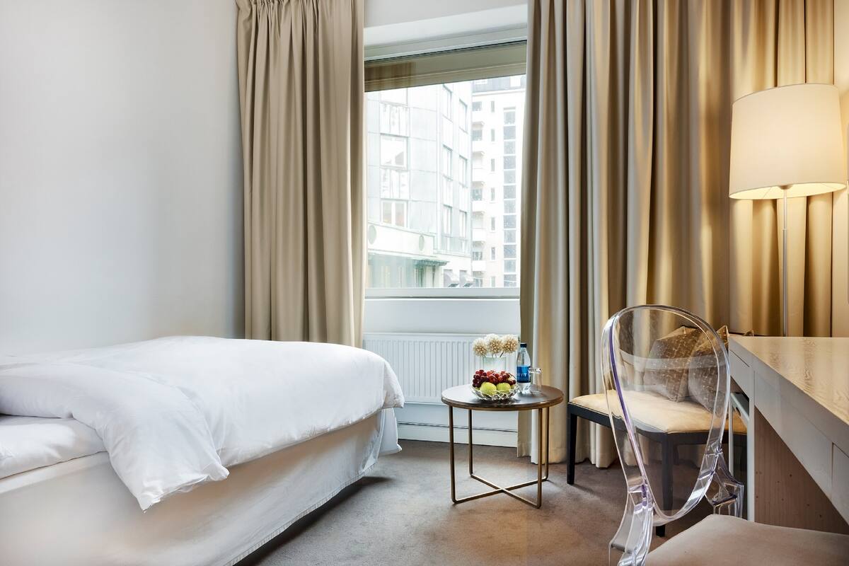 A hotel room with a bed, a small table with a fruit bowl, a chair, and a lamp.