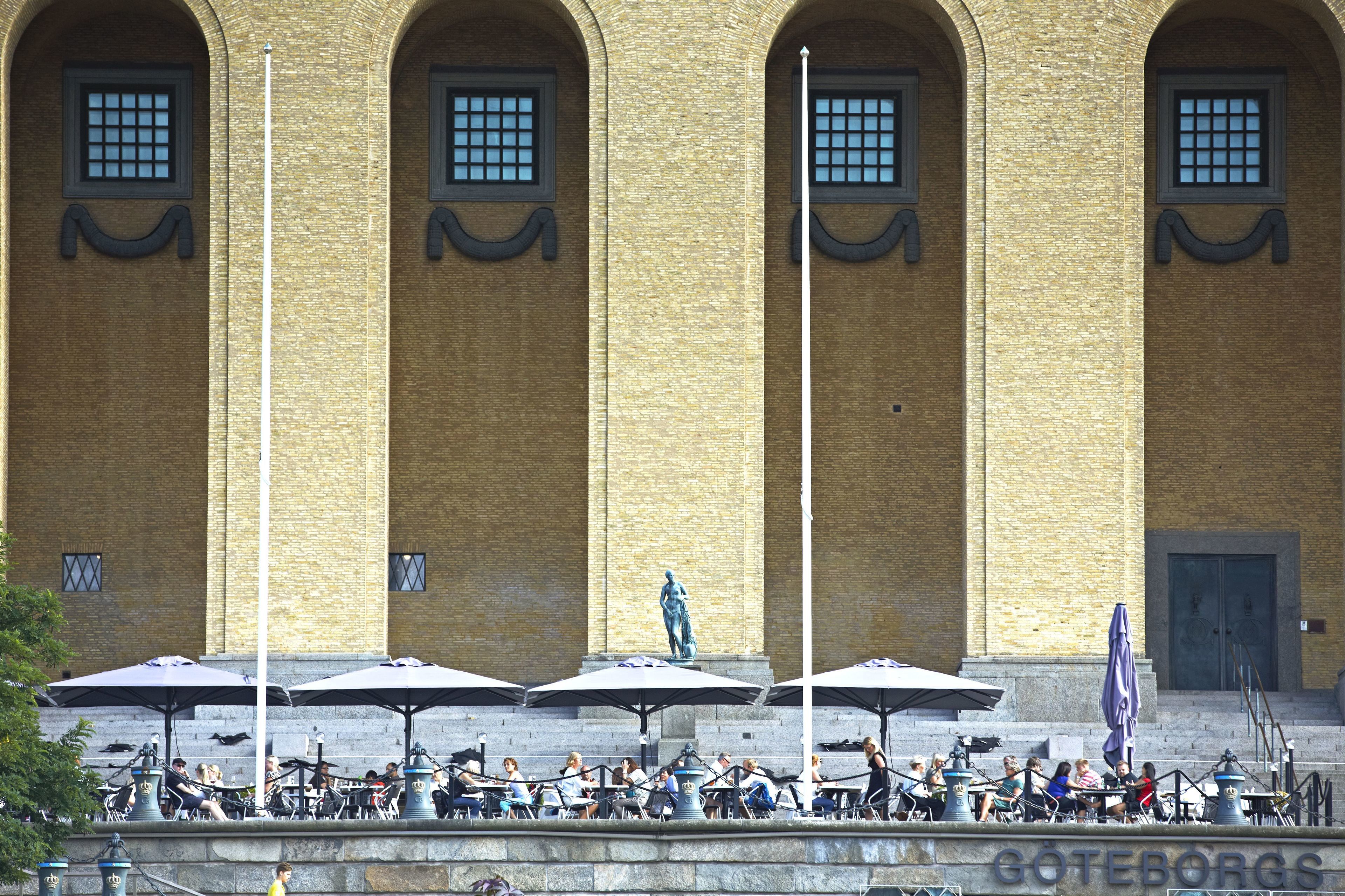 A building with arched windows and a statue in front. There are tables and umbrellas under the windows.
