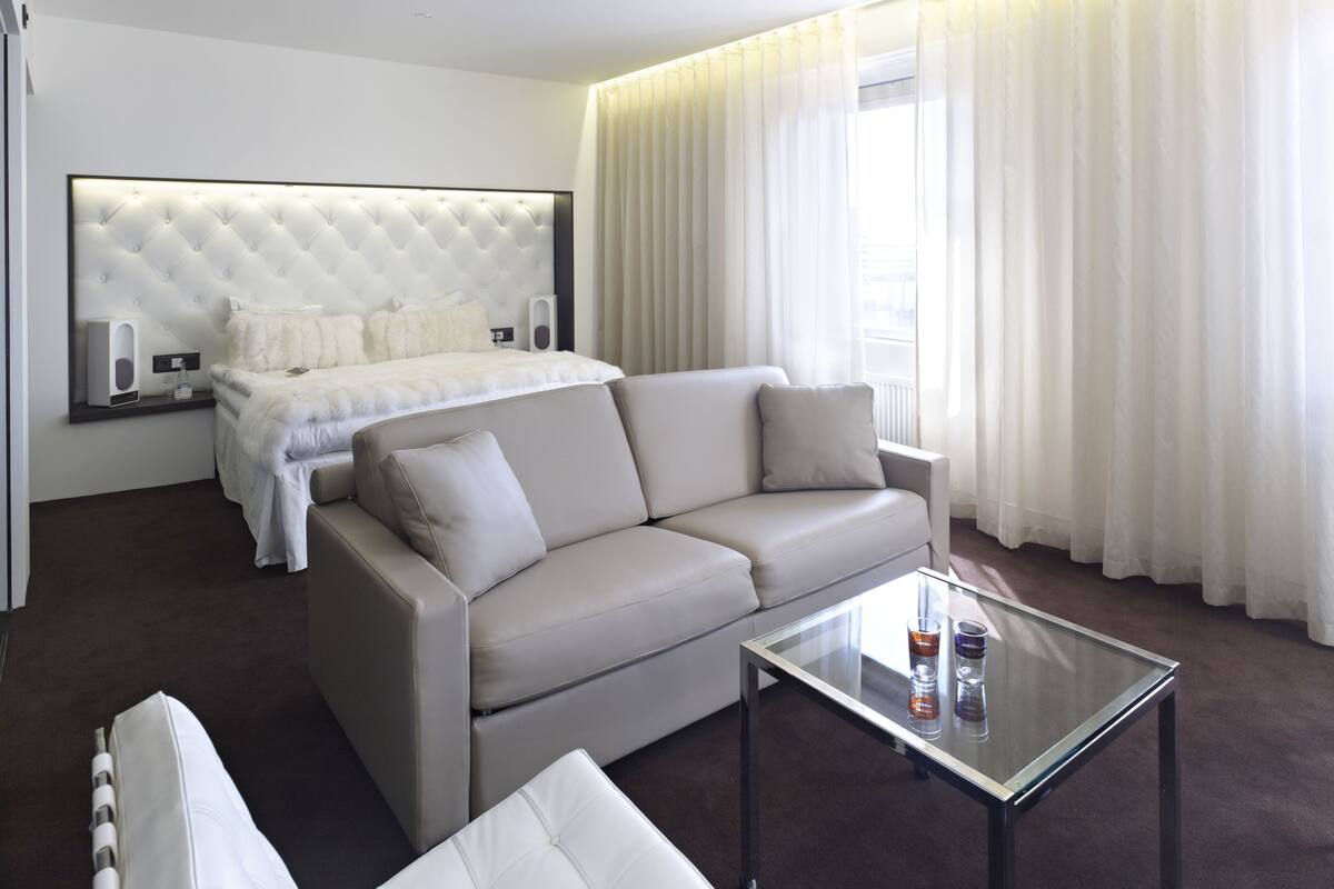 A modern hotel room with a large bed, a sofa, a glass coffee table, and a large window with curtains.