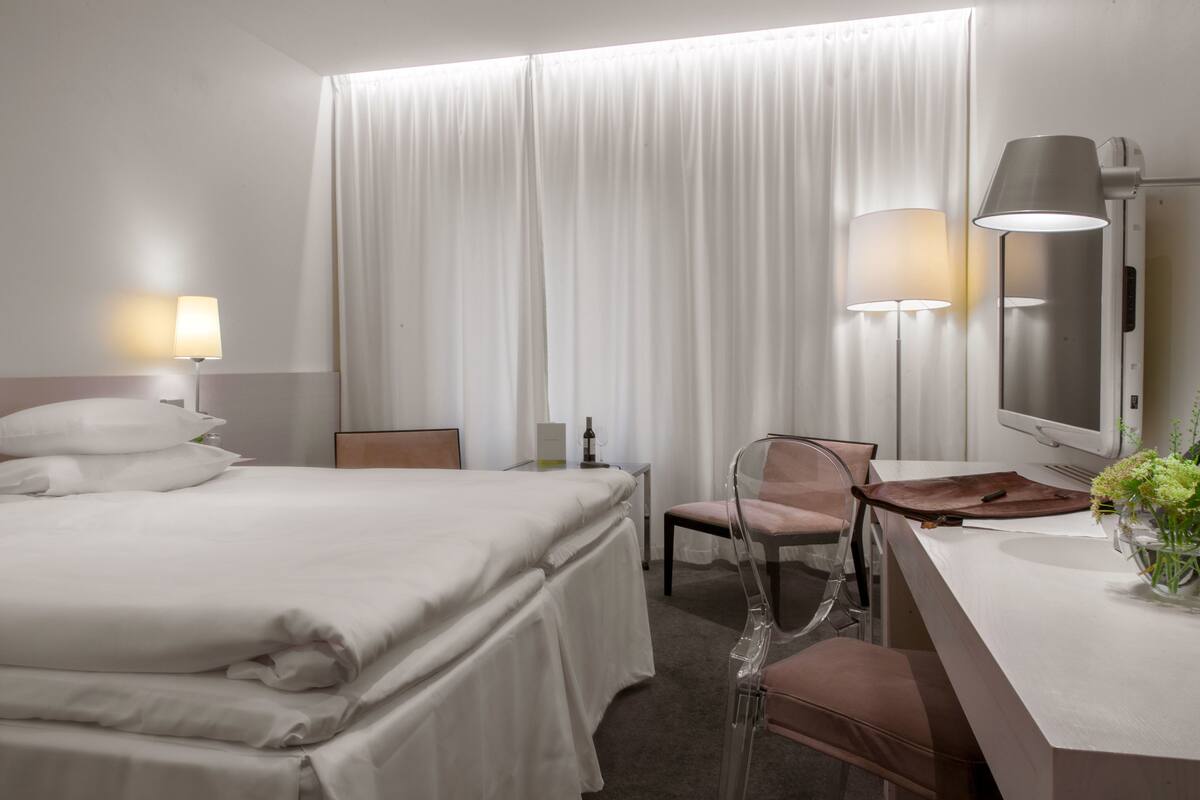 A hotel room with a bed, a desk, a chair, a lamp, a mirror, and a vase with flowers.
