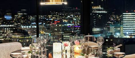 Mesa de comedor dispuesta con vasos, platos y una vela, con vista a un paisaje urbano nocturno.