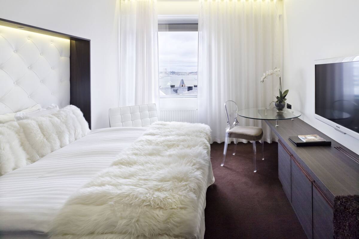 A modern hotel room with a large bed, a glass desk, a television, and a view of the city through the window.