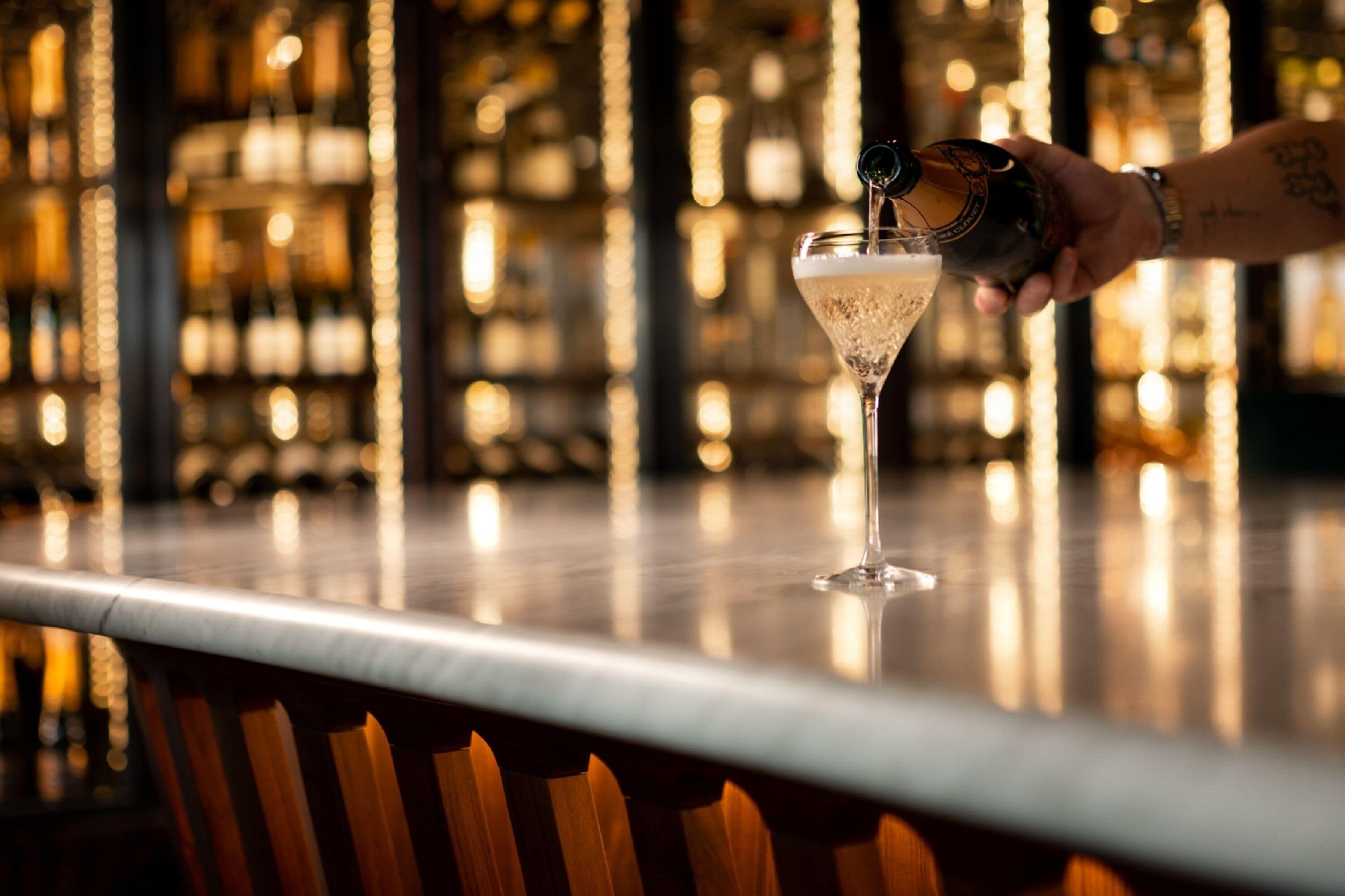 A hand pouring a drink into a martini glass on a bar counter with a blurred background of bottles and lights.