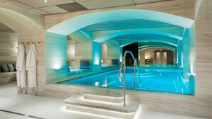 A modern indoor swimming pool with clear glass walls, a central staircase, and a seating area with a sofa and pillows. - Hotel Riverton (Gothenburg)