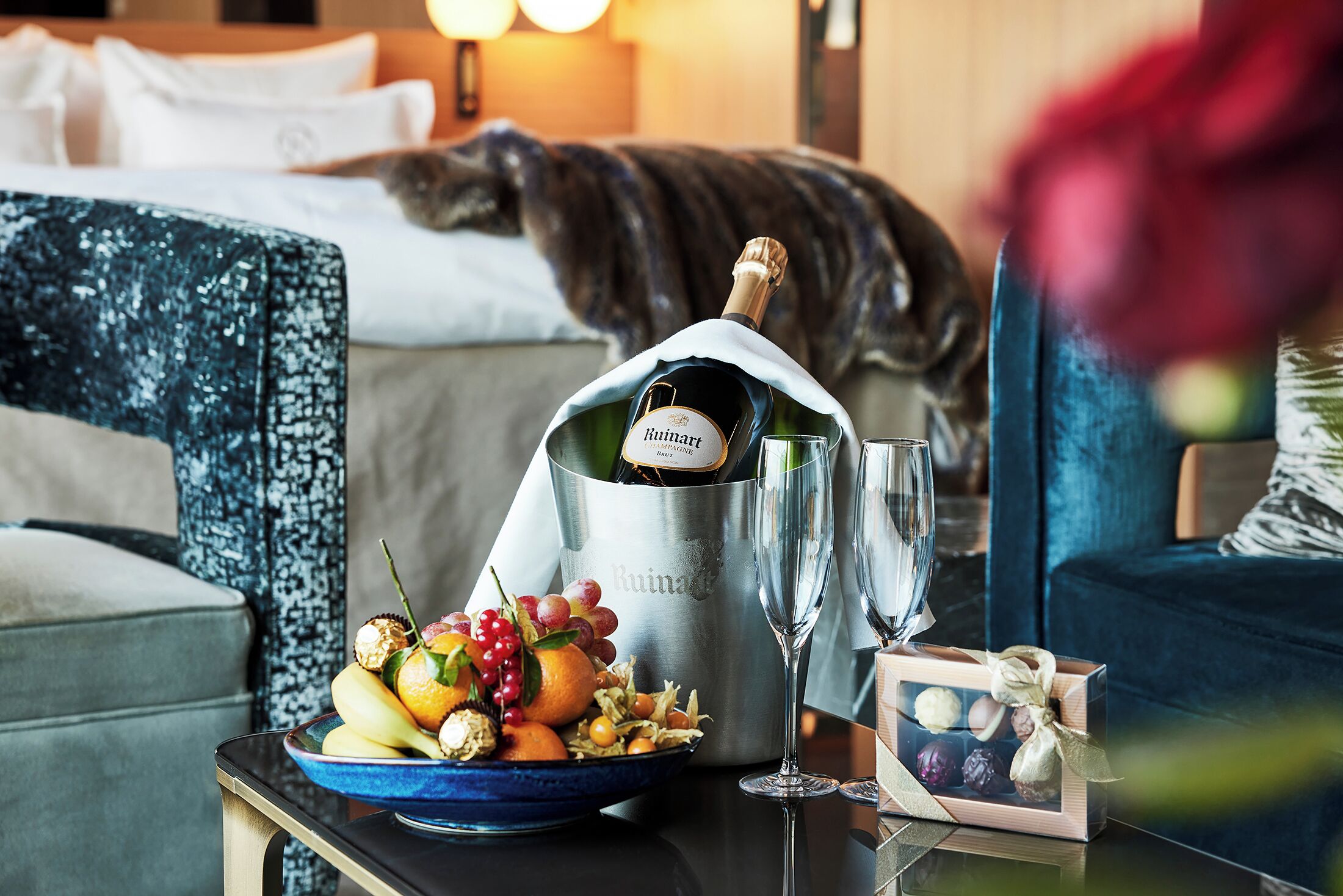 Quarto de hotel com uma cama, uma mesa de cabeceira com uma garrafa de champanhe Ruinart, uma tigela de frutas e um copo.