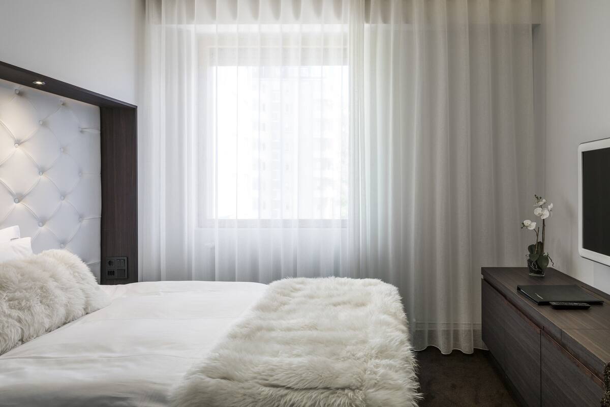 A bedroom with a tufted headboard, a fluffy white bench, a bedside table with a vase of white flowers, and a TV on a wooden console.