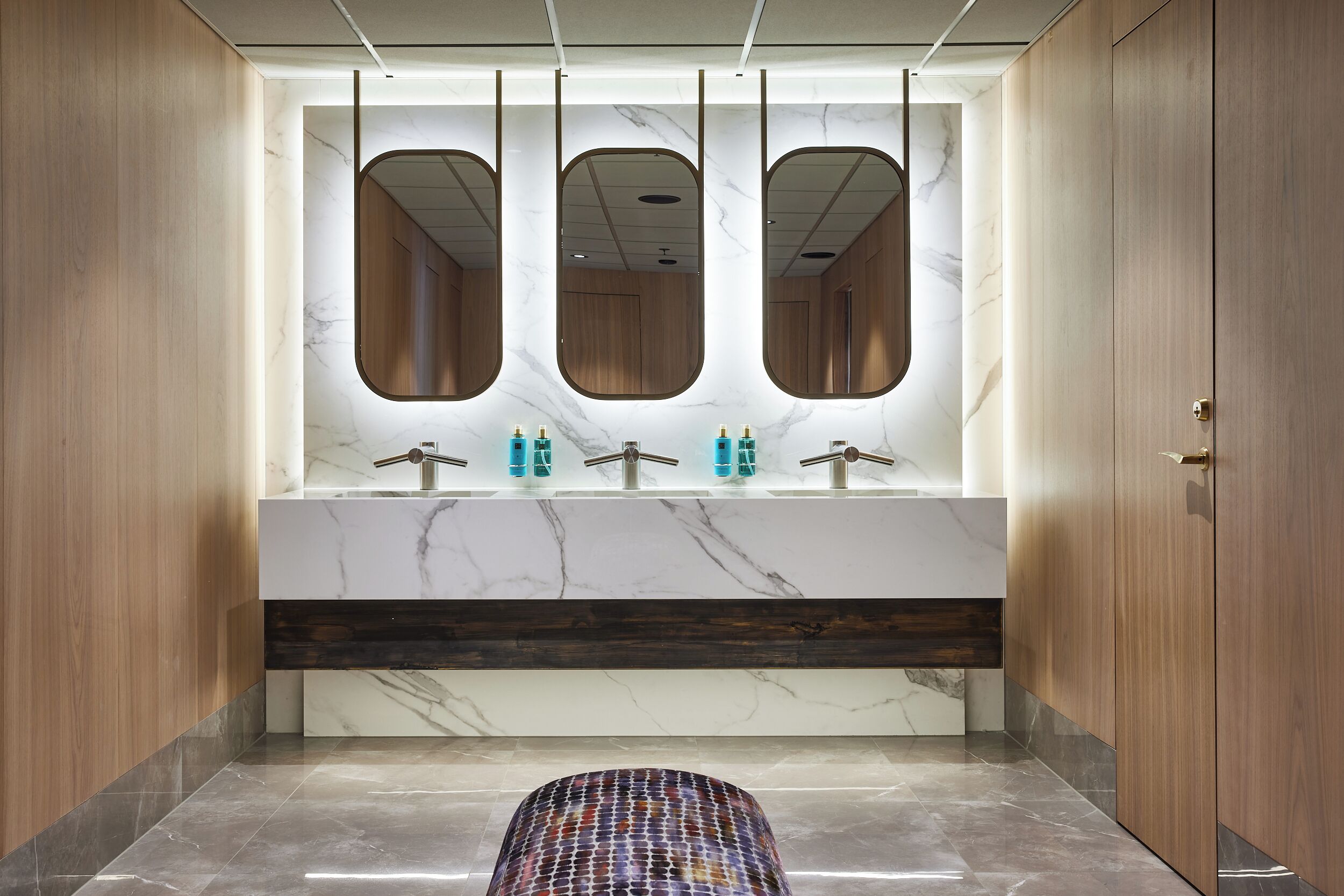 A modern bathroom with a marble vanity, three mirrors, and a patterned stool.