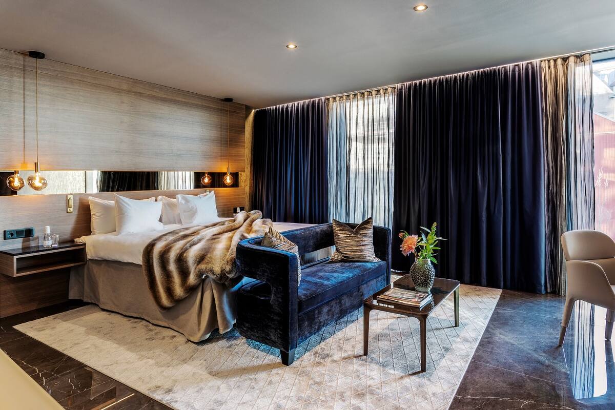 A modern hotel room with a bed, a dark blue sofa, a small table with a vase, and a large window with curtains.