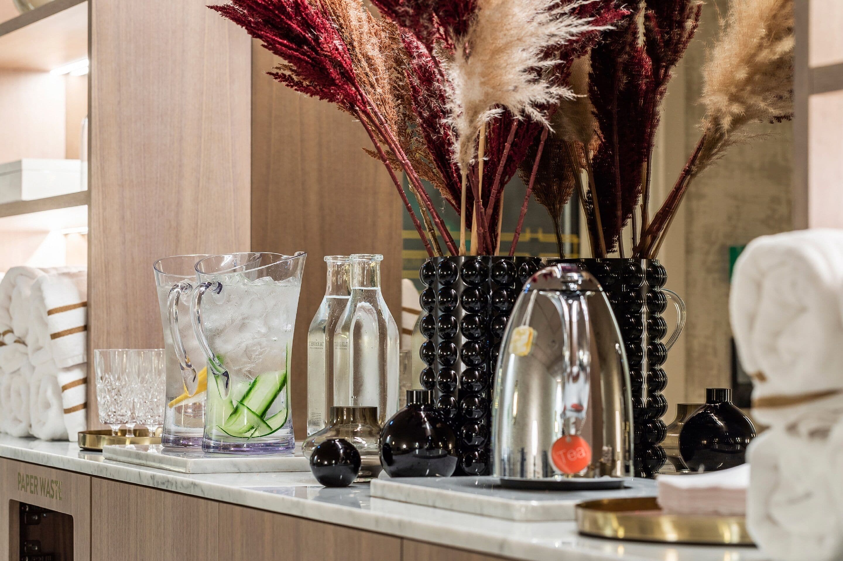 A bar area with various glassware, a pitcher with ice and cucumber slices, and decorative dried plants.