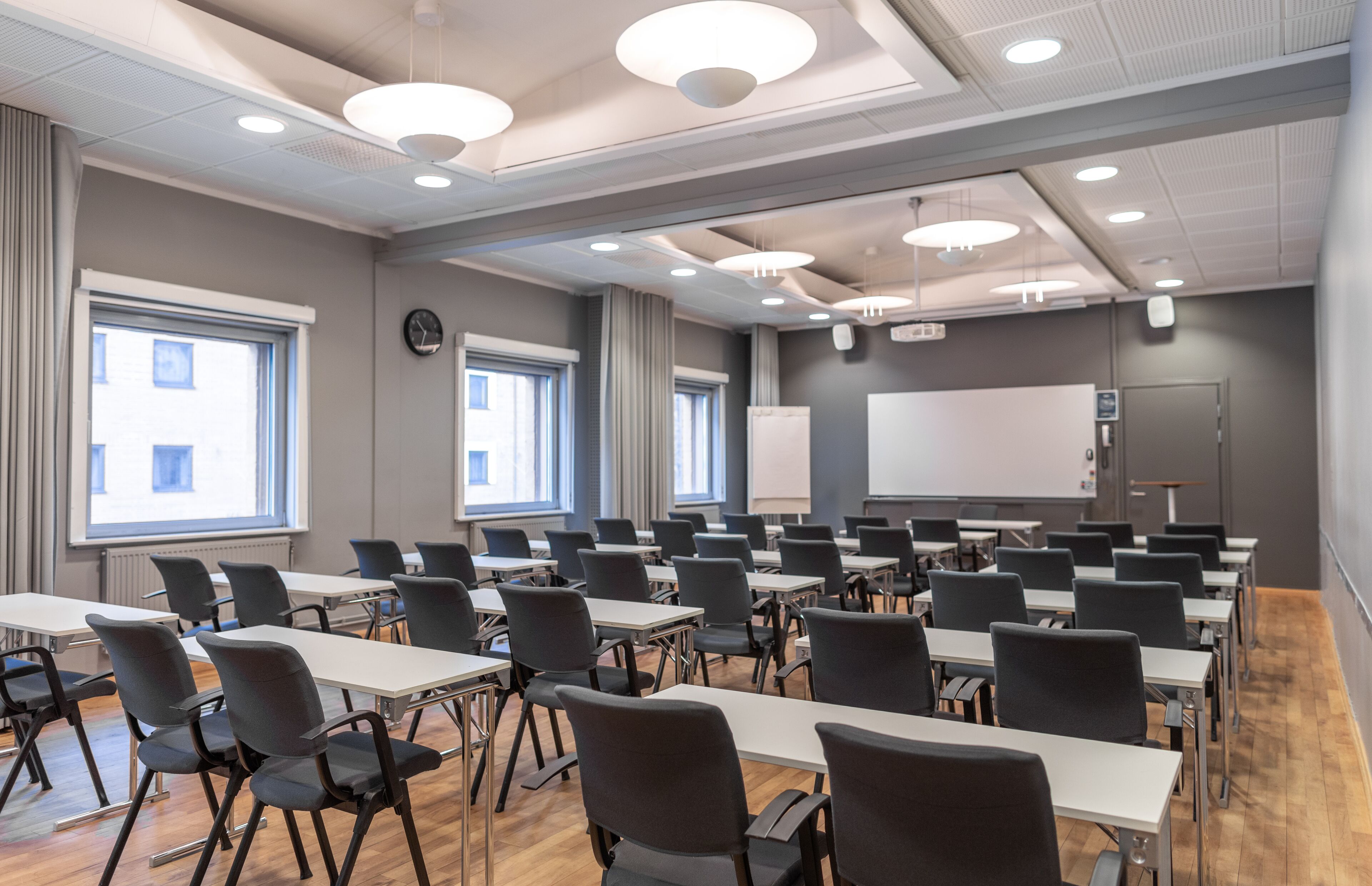 A conference room with rows of tables and chairs, large windows, a whiteboard, and a projector screen.