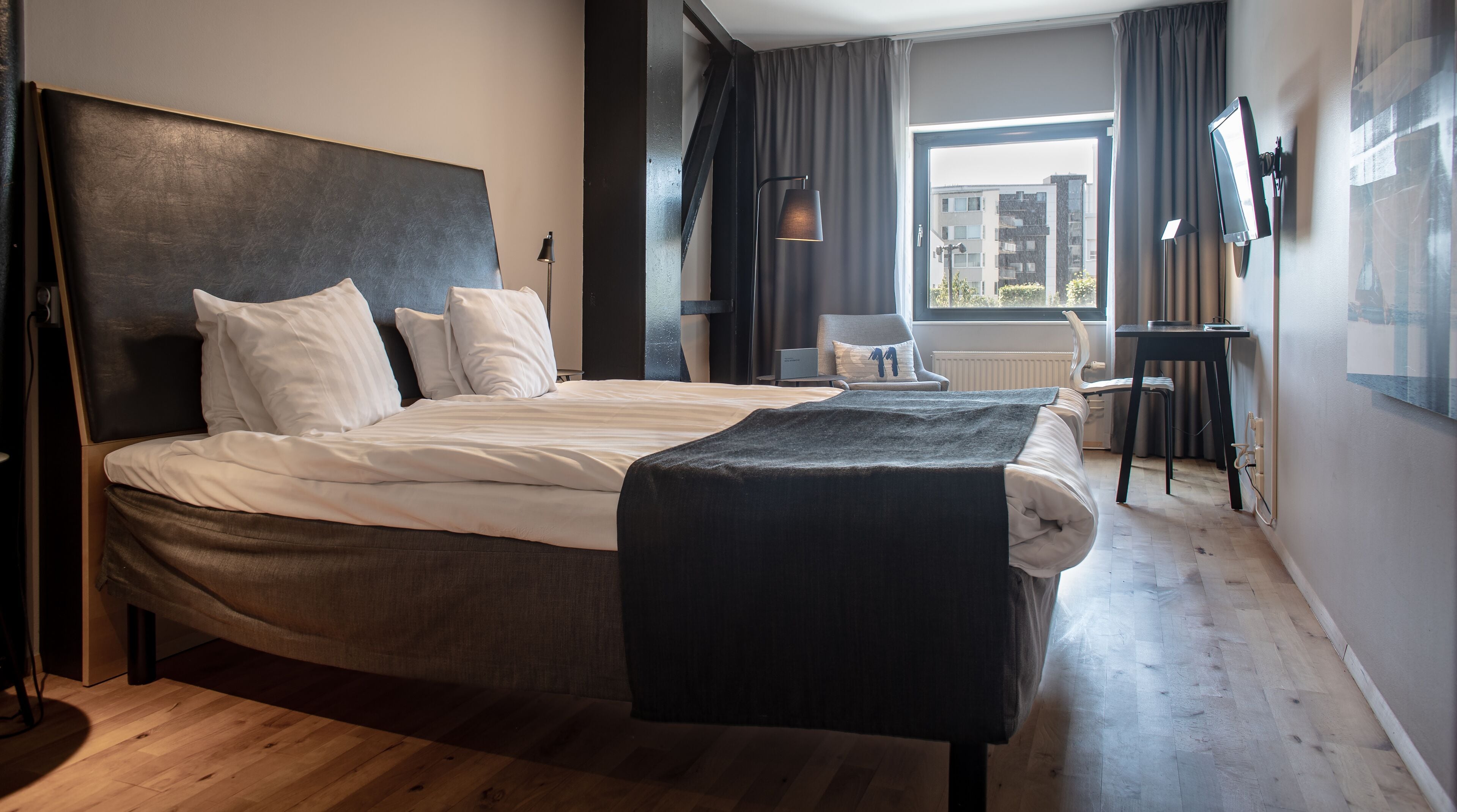 A modern hotel room with a large bed, a desk, and a view of the city through the window.