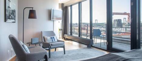 A modern hotel room with a large window, a flat-screen TV, a sofa, a chair, and a small table.