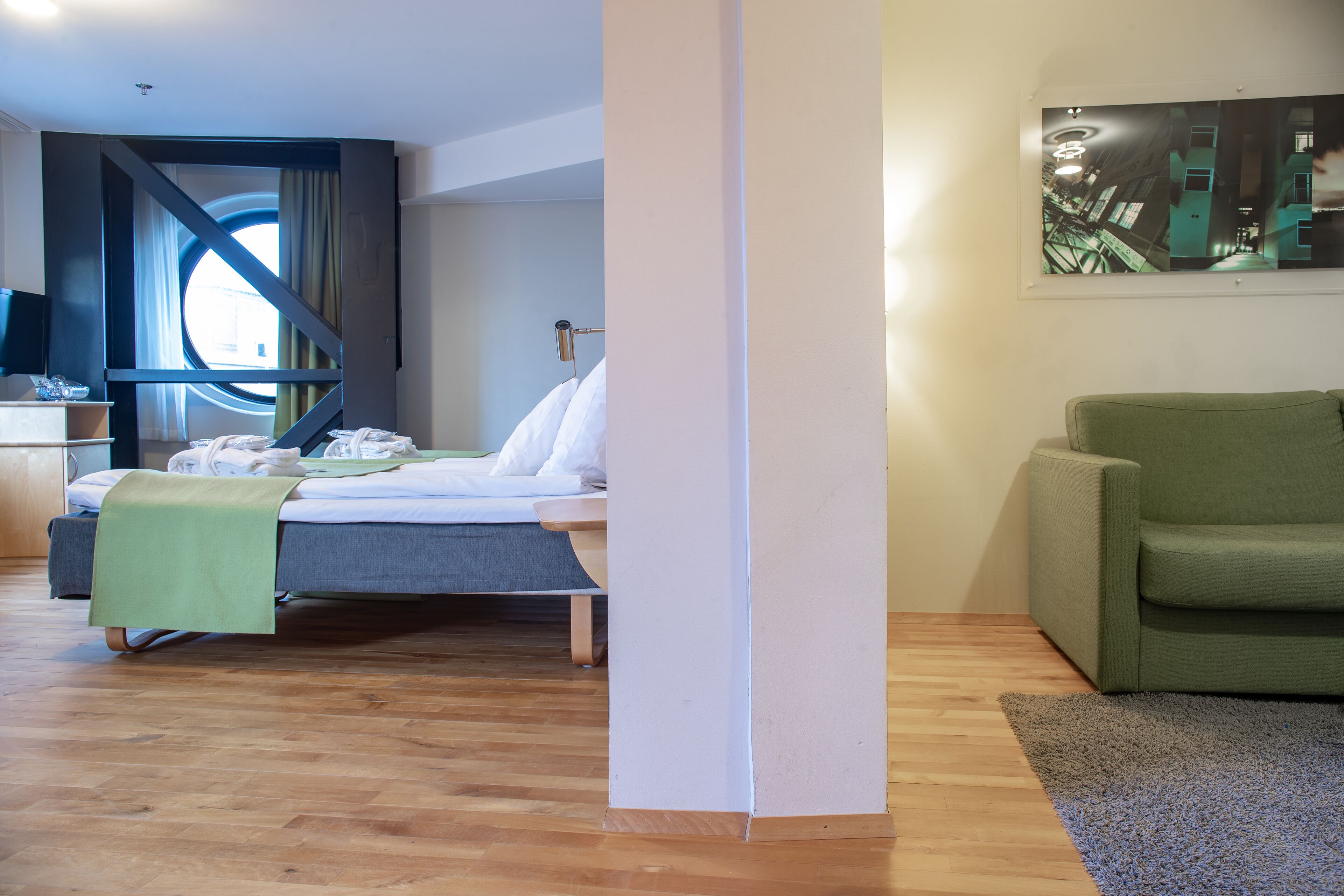 A modern hotel room with a bed, a green sofa, a painting on the wall, and a window with curtains.
