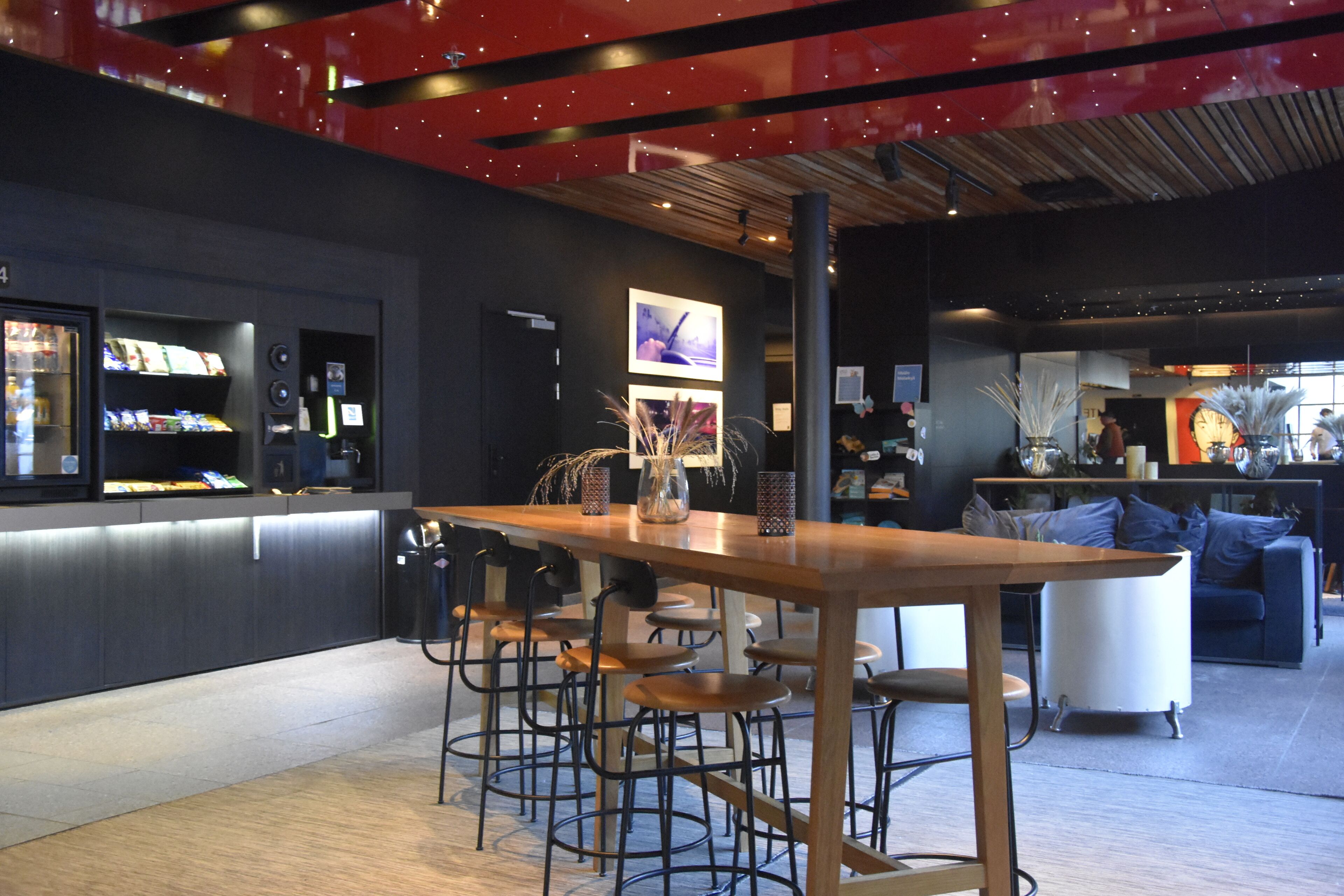 A modern bar area with a long wooden table, multiple bar stools, and a counter with shelves of books and magazines.