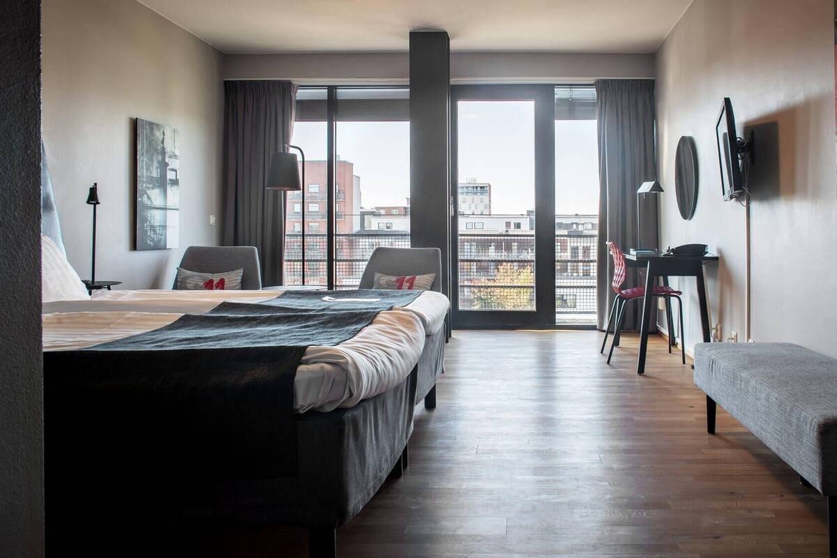 A modern hotel room with a large bed, a desk, a chair, and a balcony with a city view.