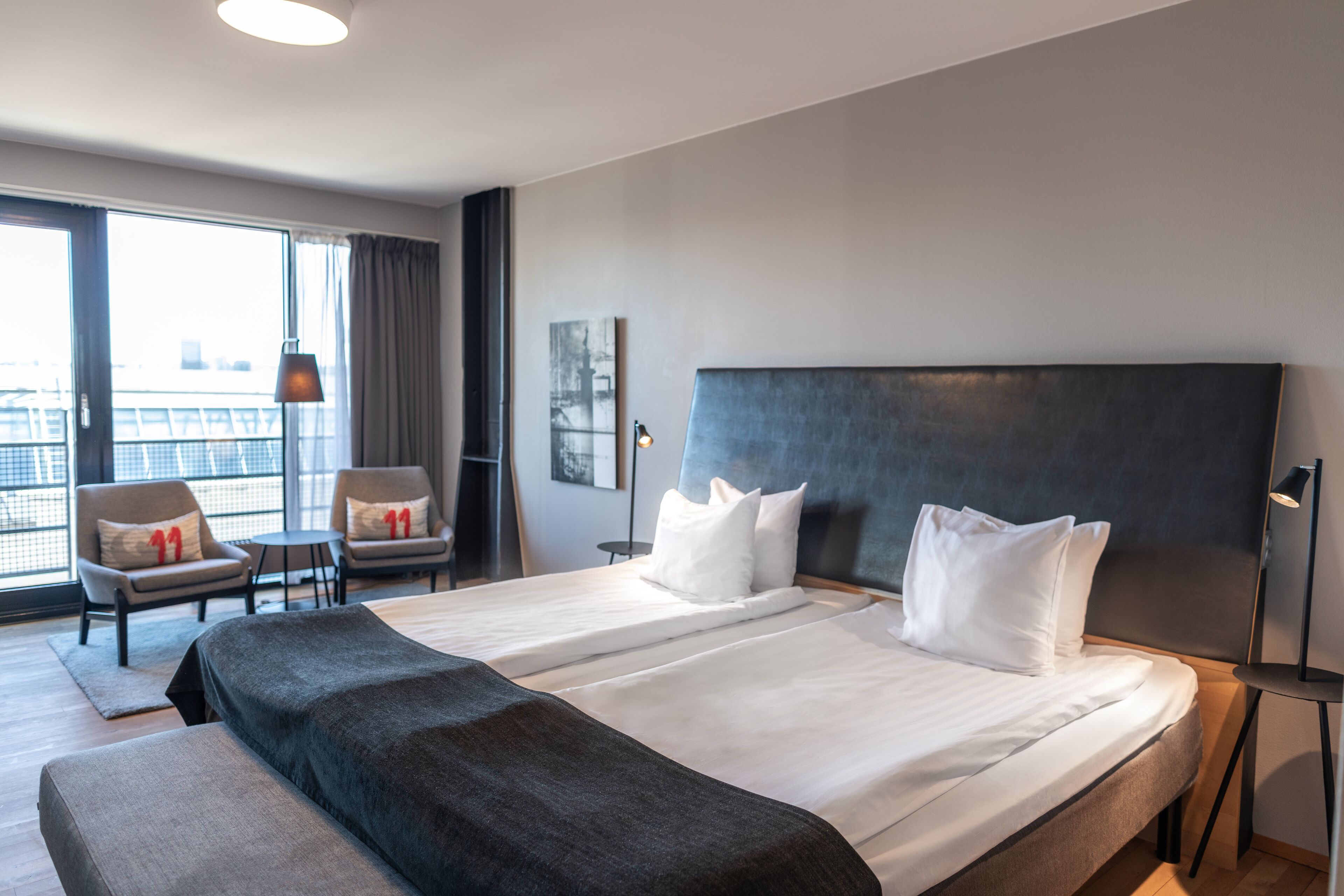 Habitació d'hotel moderna amb un llit gran, dues butaques, una taula petita i vista a la ciutat a través de grans finestres.