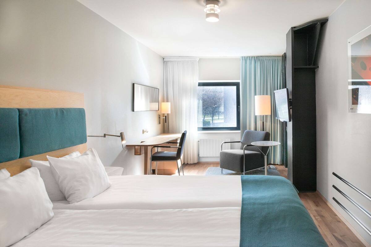 A hotel room with two beds, a desk, a chair, a TV, and a window with curtains.