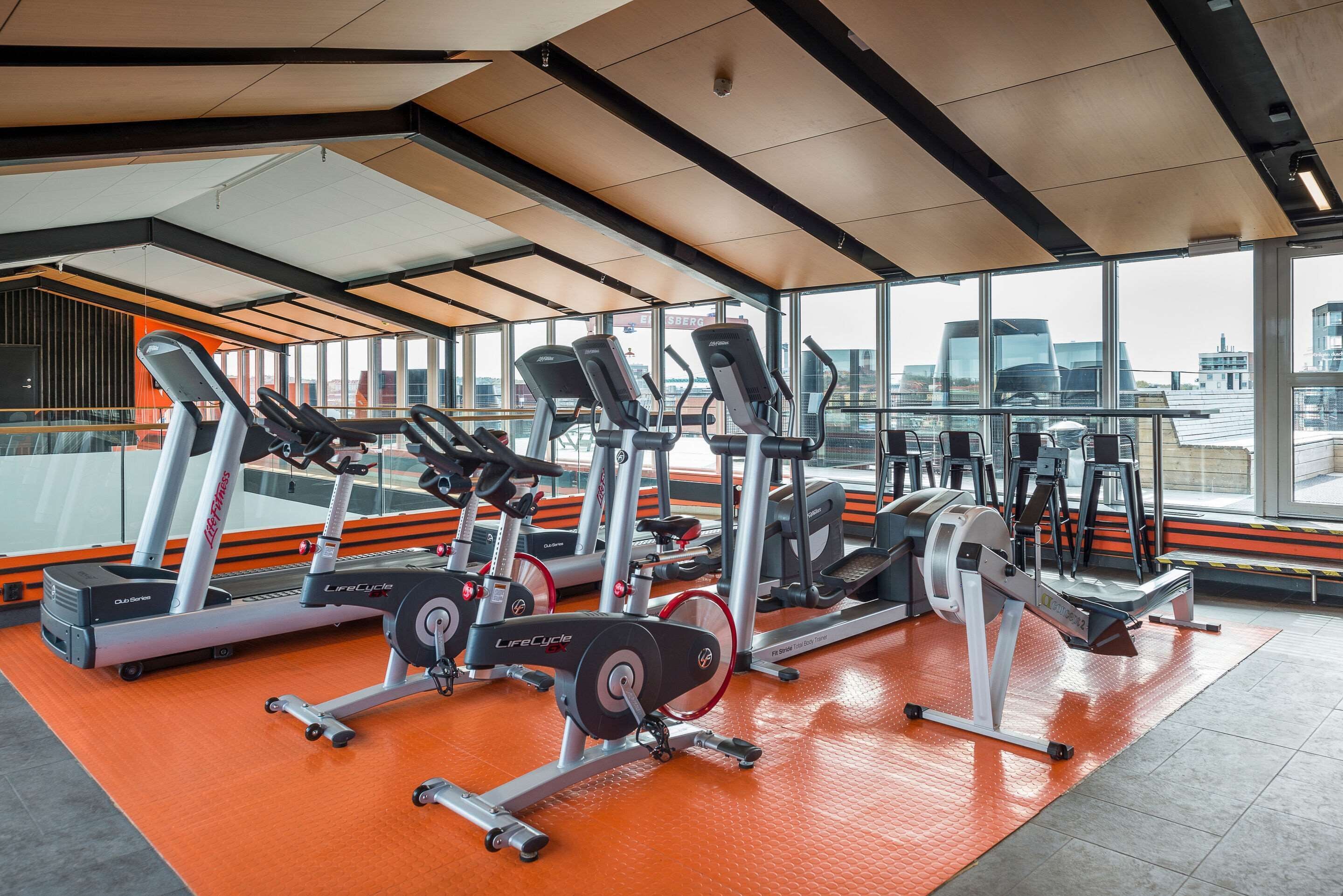 Une salle de sport équipée de divers appareils d’exercice, dotée de grandes fenêtres et offrant une vue sur le paysage urbain.