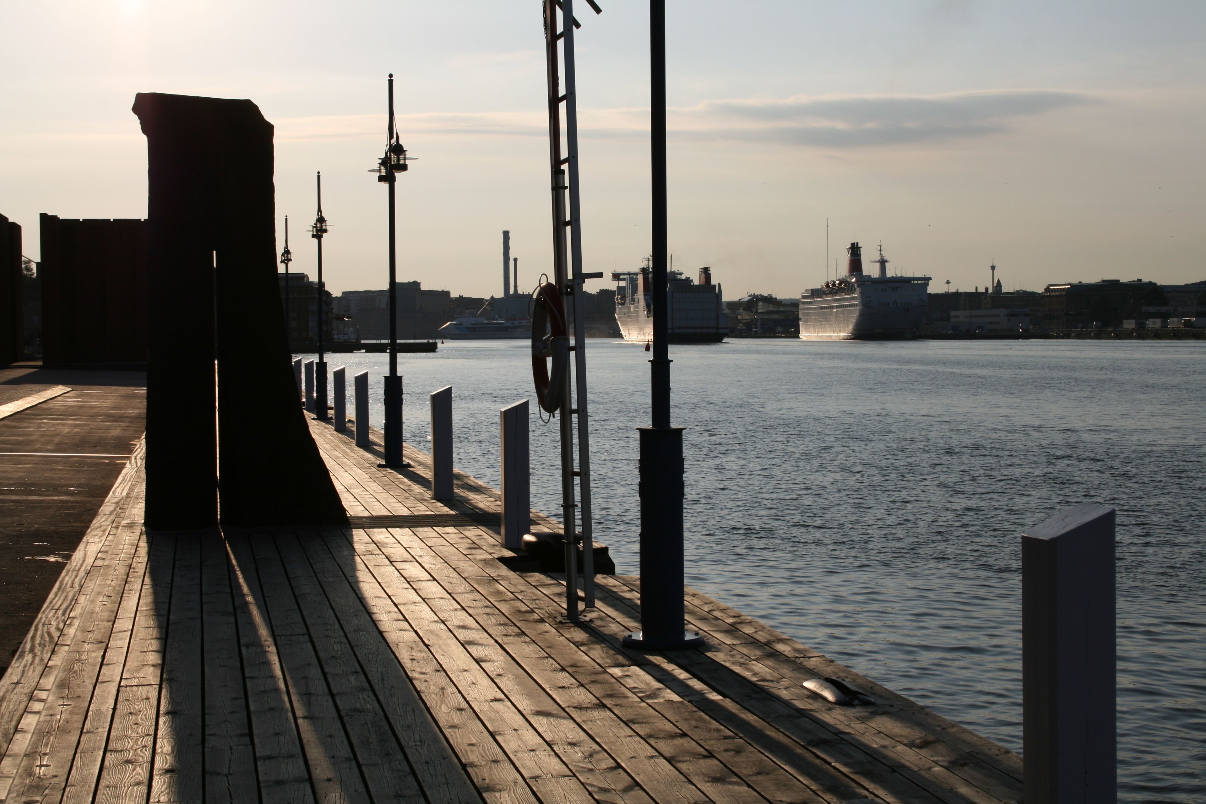 A wooden pier extending into calm water, with a ship docked in the background and a cityscape in the distance.