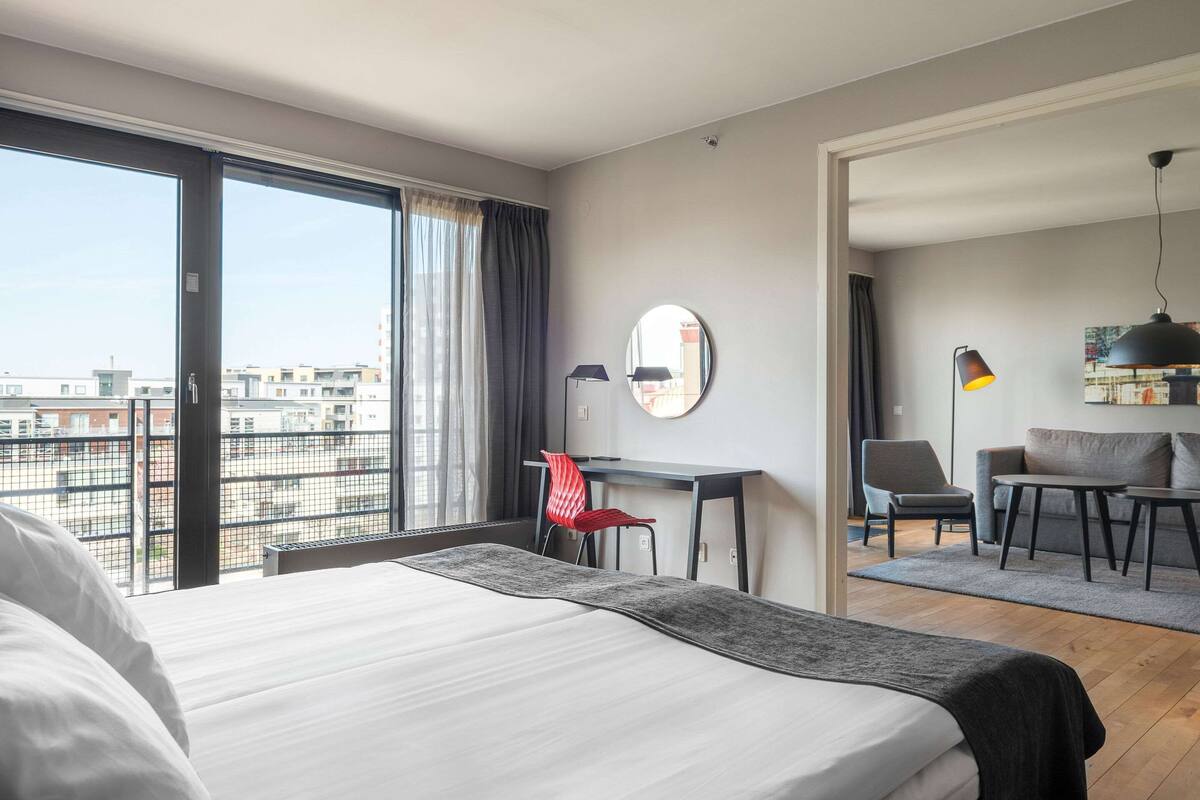 A modern hotel room with a large bed, a desk, a chair, and a view of the cityscape through large windows.