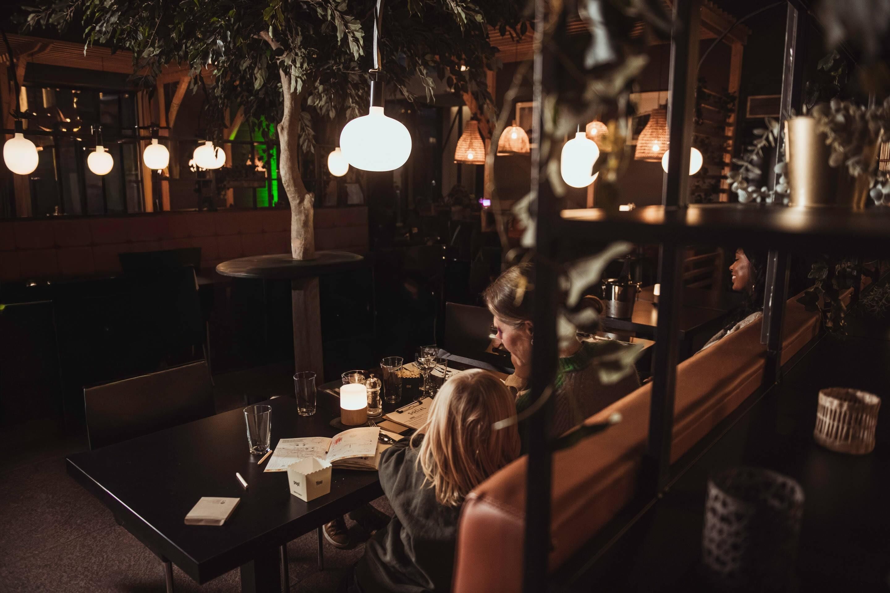 Un restaurante tenuemente iluminado con mesas y sillas, lámparas colgantes y una vista del interior a través de una ventana.