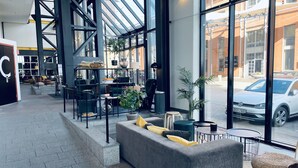 A modern interior with a grey sofa, a wicker chair, and a large window overlooking a street. - Quality Hotel 11 (Gothenburg)