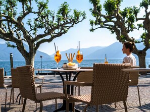 A table with two glasses of orange drink, a plate with a pastry, and a bowl of breadsticks, set outdoors with wicker chairs and a view of the sea and mountains. - Albergo Carcani (Ascona)