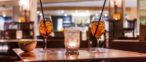 Zona de bar con dos vasos de bebidas ámbar con pajitas, un bol de aperitivos y una vela encendida sobre una mesa de madera.