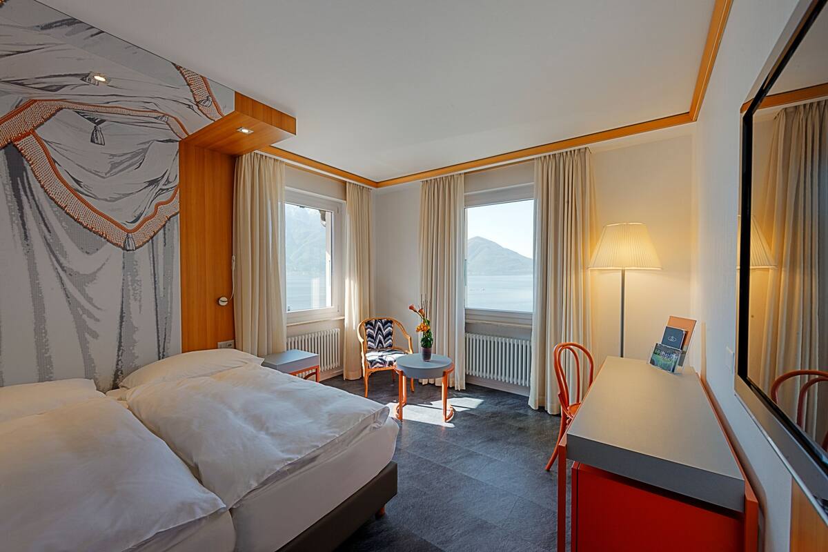 A hotel room with a bed, a small table, a chair, and a view of the mountains.