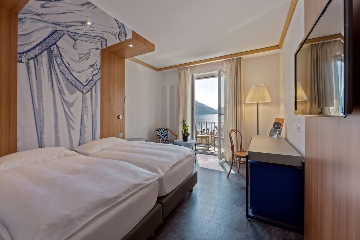 A hotel room with a bed, a desk, a chair, a lamp, and a view of the outdoors.