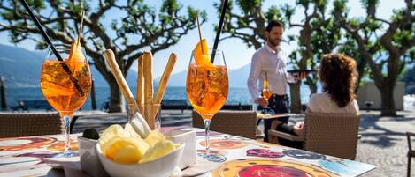 An outdoor dining area with a table set for two, featuring a bowl of fruit, two glasses of orange beverage, and breadsticks.