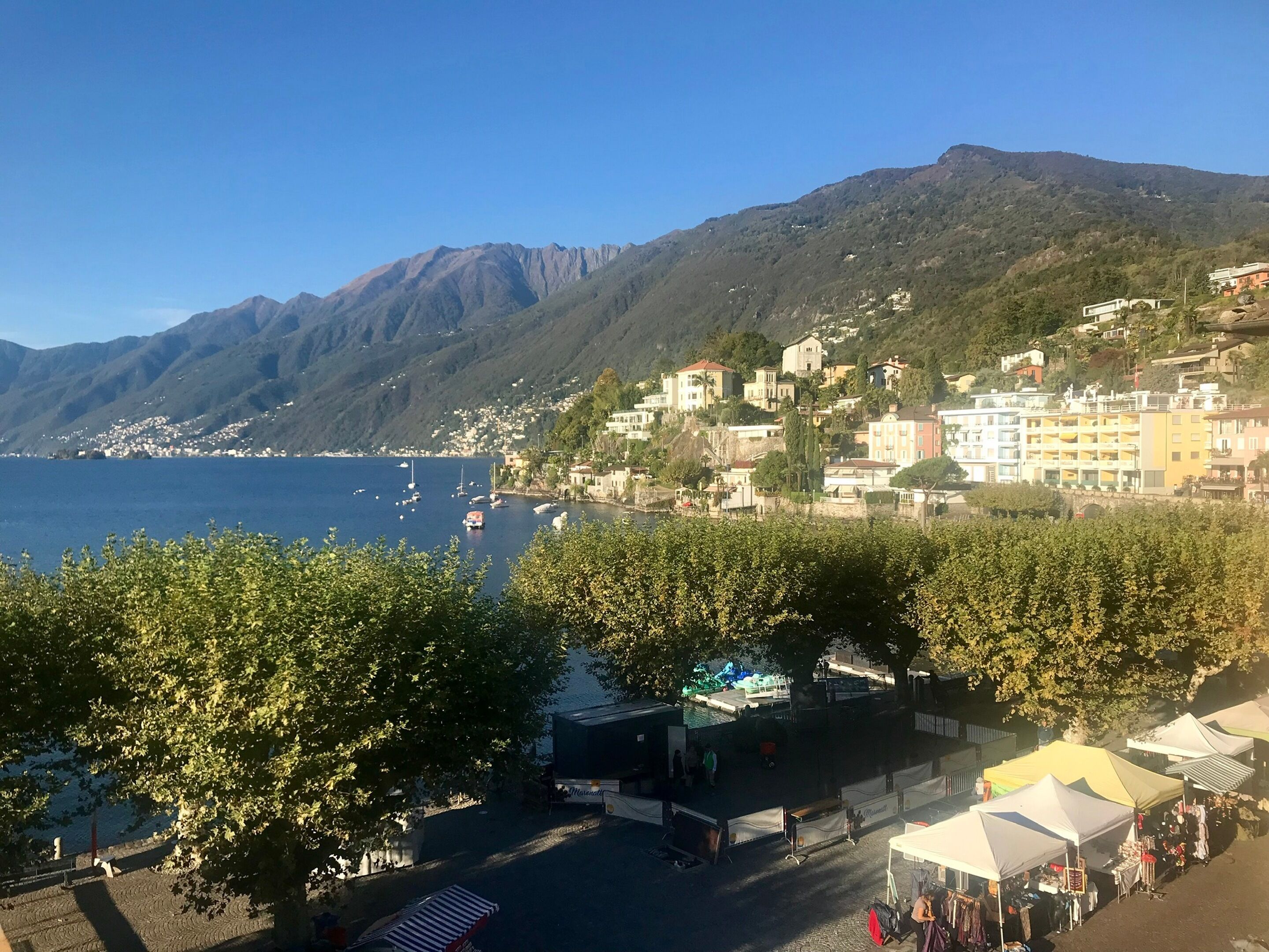 A coastal town with buildings on a hillside, a clear blue sea, and a market area with tents.