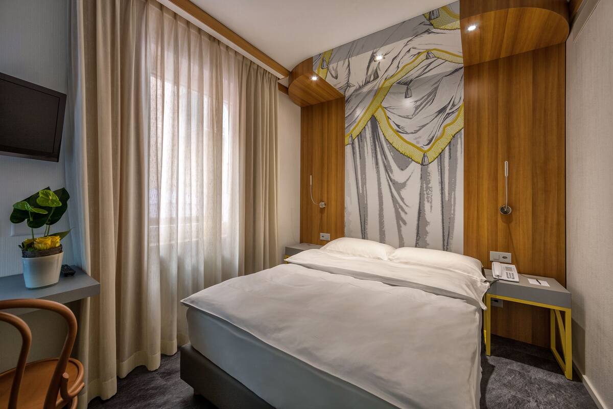 A hotel room with a bed, a desk with a plant, a telephone, and a wall with a decorative design.