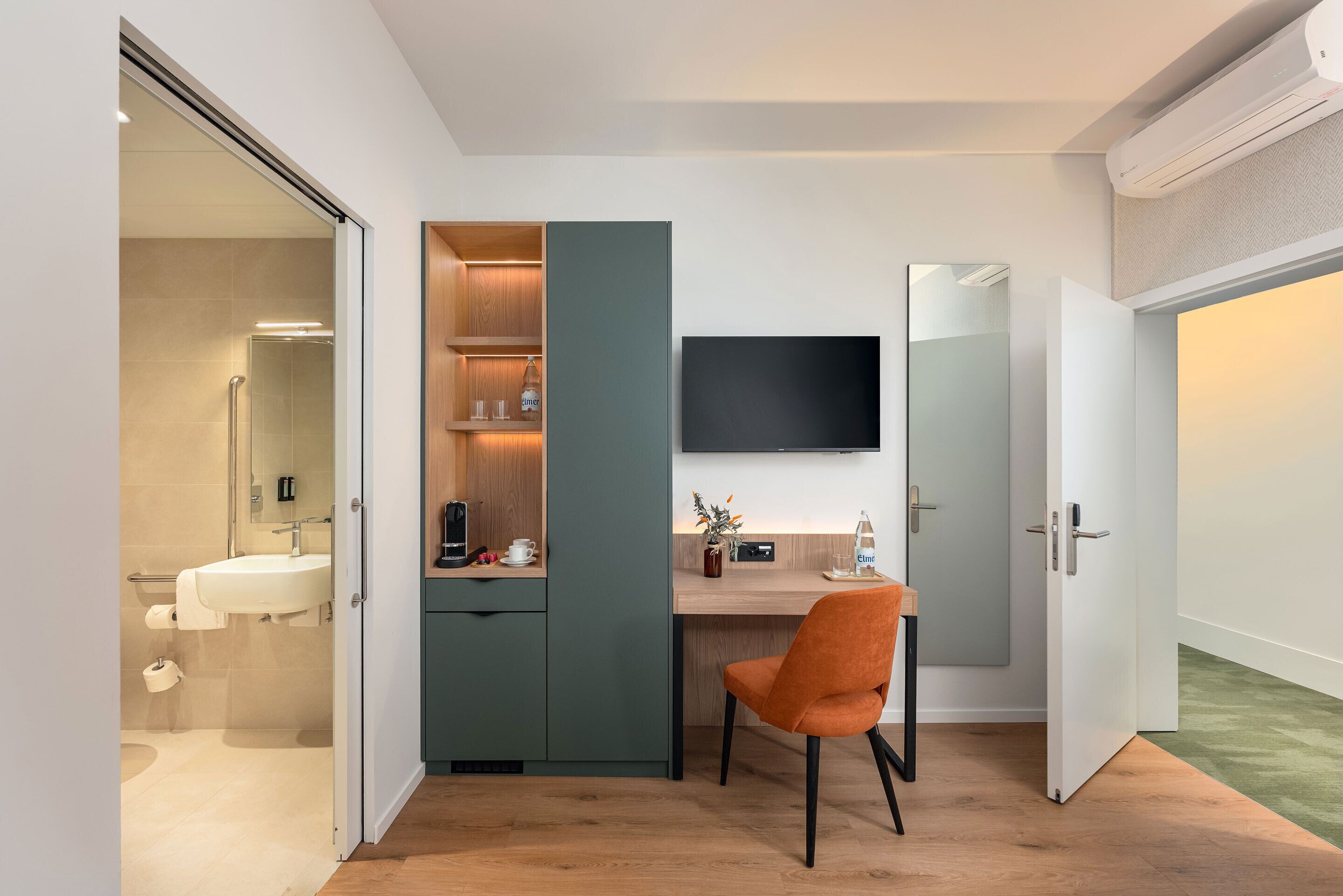 A modern bathroom with a shower, toilet, and sink. There is a wooden cabinet with a television above it and a chair in the room.